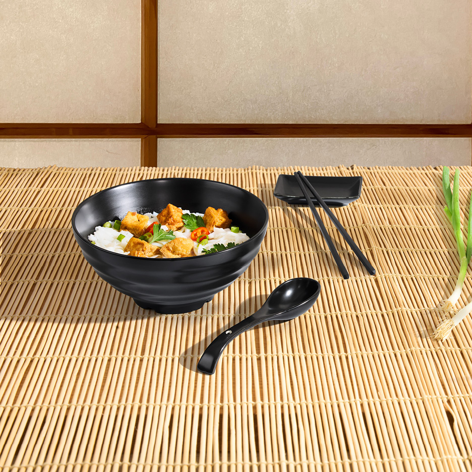 Svart skål med ris och kyckling, svarta ätpinnar och sked på en bambumatta.