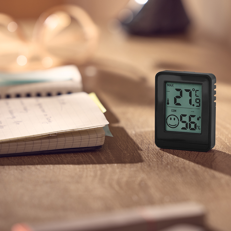 En digital termometer och hygrometer på ett träbord bredvid en anteckningsbok.