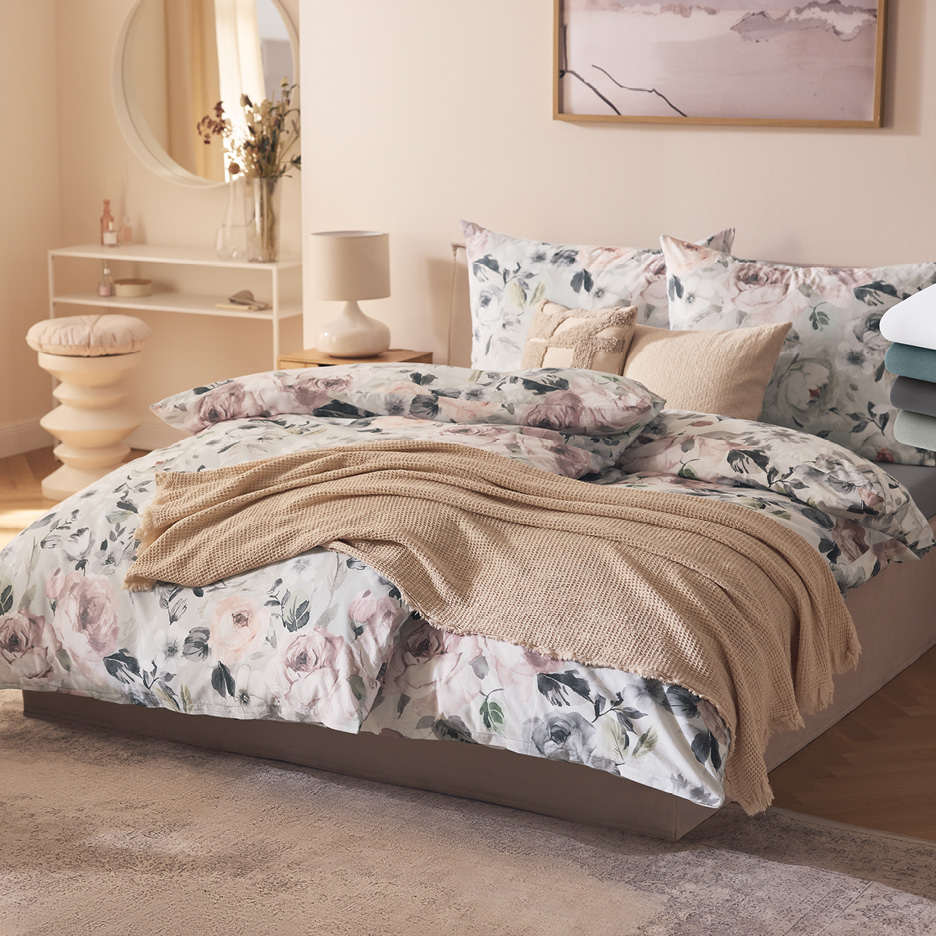 Sovrum med säng klädd i blommiga sängkläder, prydnadskuddar och en beige pläd.