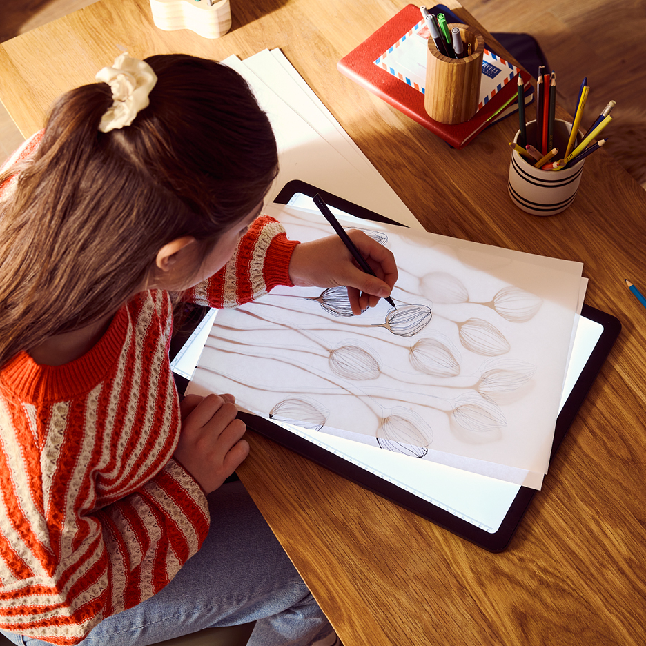 En flicka ritar på ett ritbord med en penna, med papper och pennor i närheten.