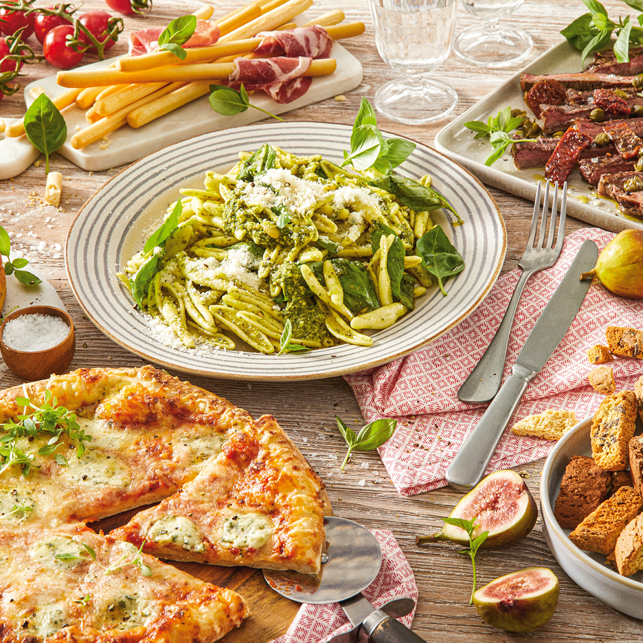En festlig måltid med pesto-pasta, pizza, charkuterier, brödpinnar och fikon på ett träbord.