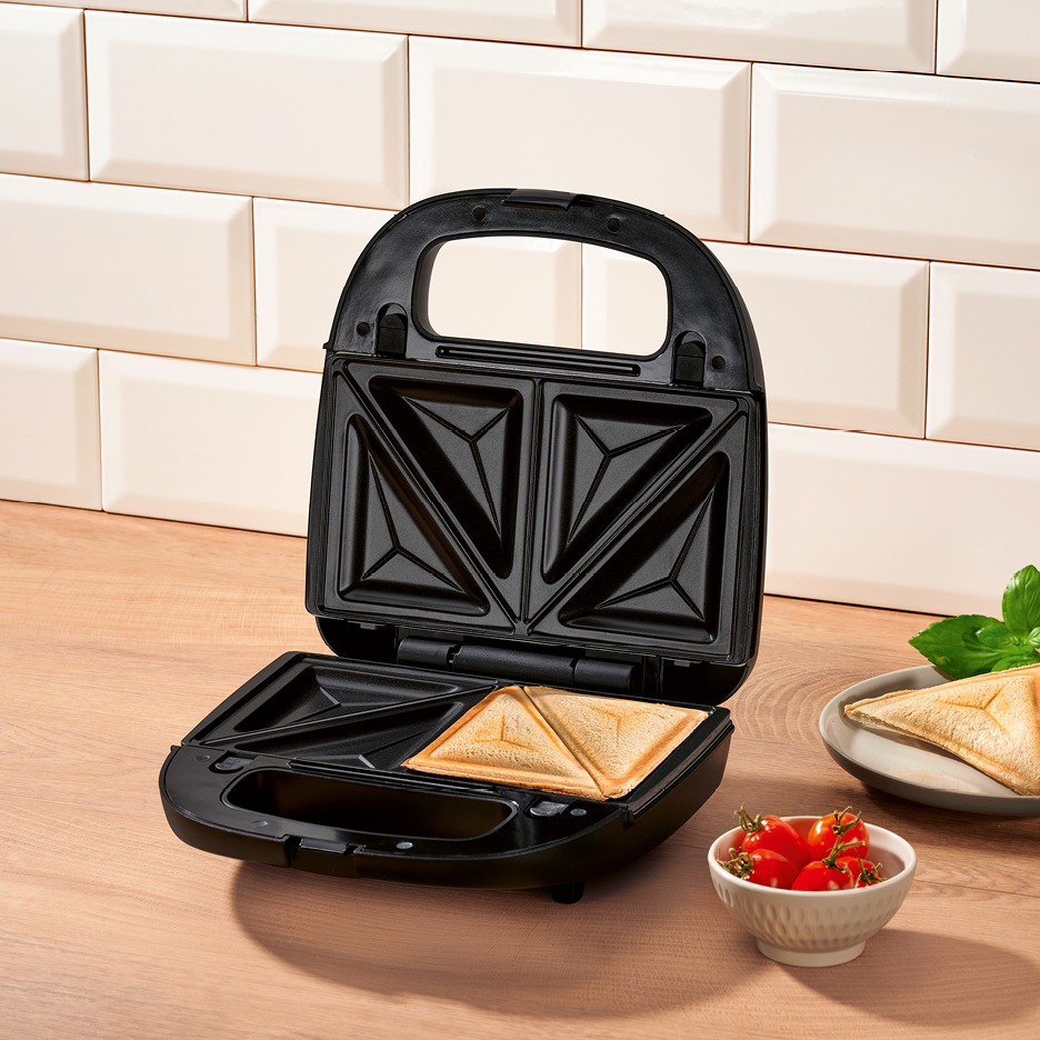 Svart smörgåsgrill med en färdig smörgås, körsbärstomater och basilika på en träbänk.