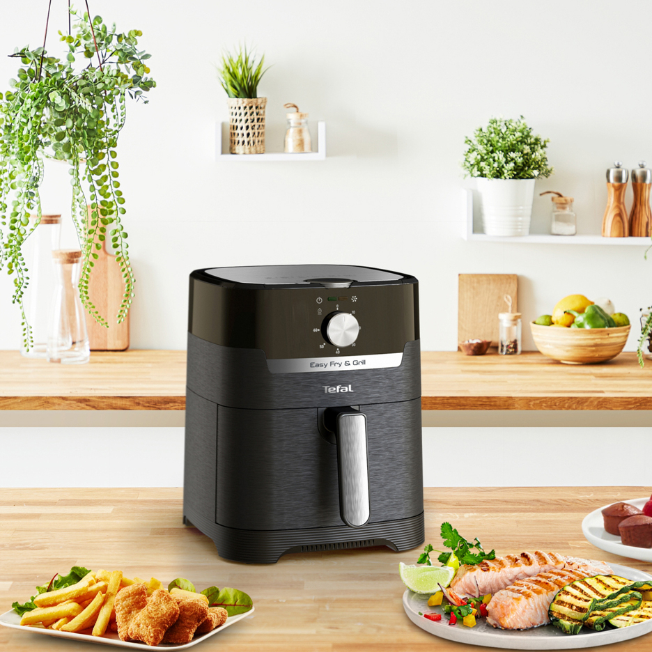 Svart airfryer på en köksbänk med tallrikar med mat som pommes frites och lax.