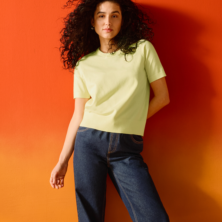Kvinna i ljusgrön t-shirt och mörkblå jeans poserar mot en orange vägg.