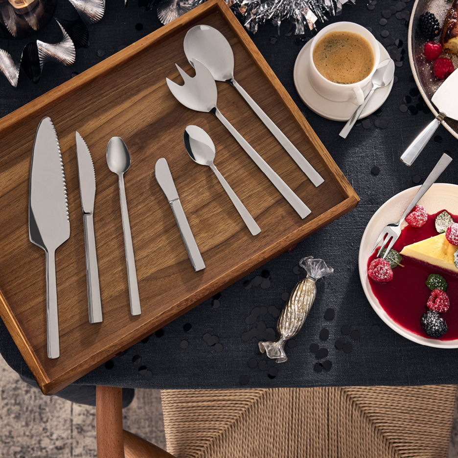 Serveringsbestick i rostfritt stål på en träbricka, med kaffe och tårta på ett festligt dukat bord.