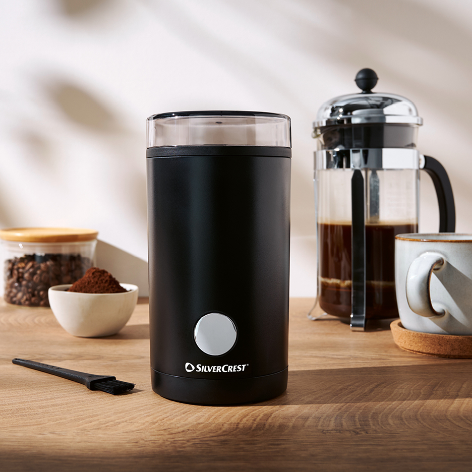 Svart kaffekvarn med en fransk press, kaffebönor och malt kaffe på ett träbord.