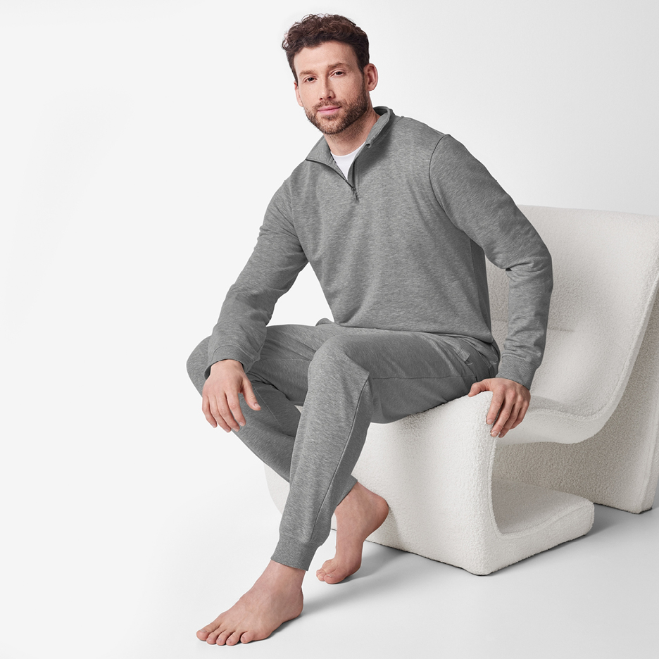 Man i grå mysdress med tröja med dragkedja och matchande byxor, sittande på en vit stol.