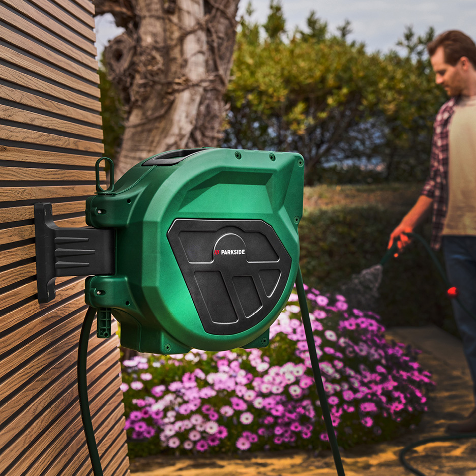 Grön väggmonterad slangvinda med en man som vattnar blommor i bakgrunden.