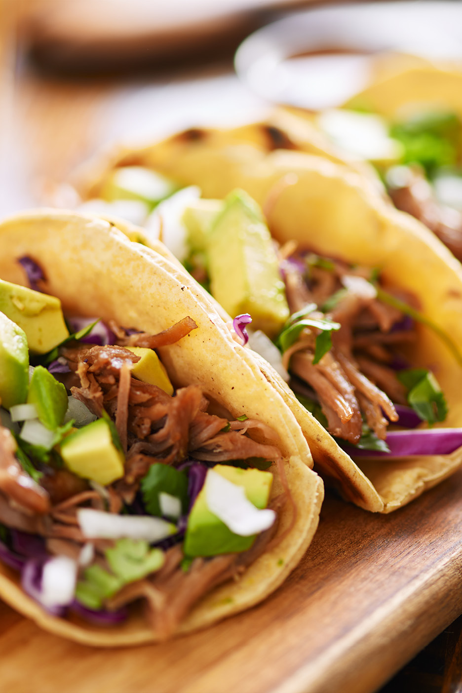 Närbild på tre tacos med pulled pork, avokado, rödkål och lök på en träskärbräda.