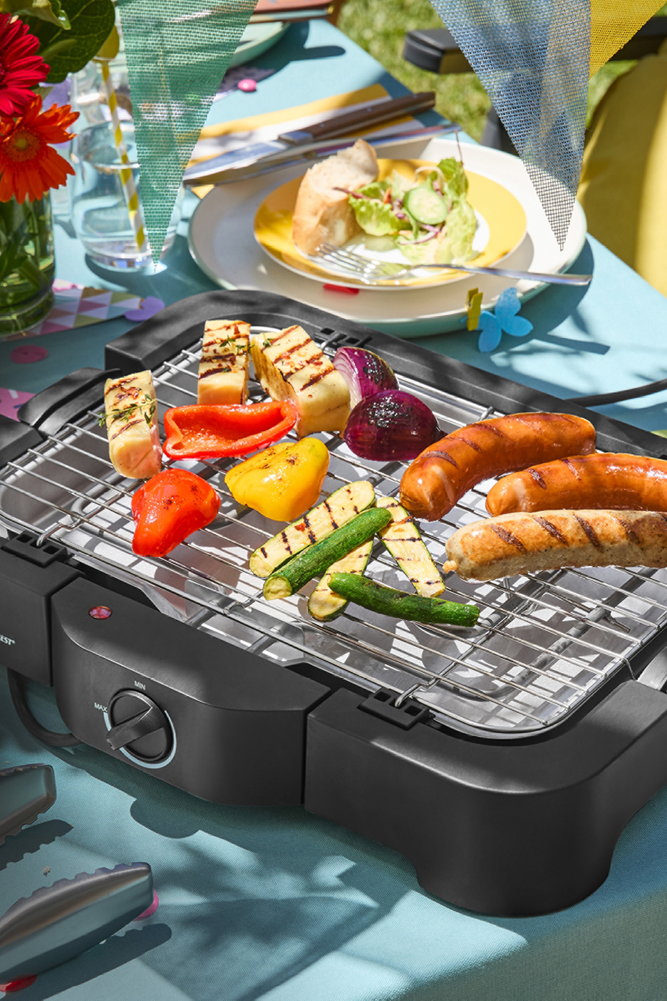 Elektrisk grill med korv, halloumi, paprika, lök och zucchini på ett dukat bord utomhus.