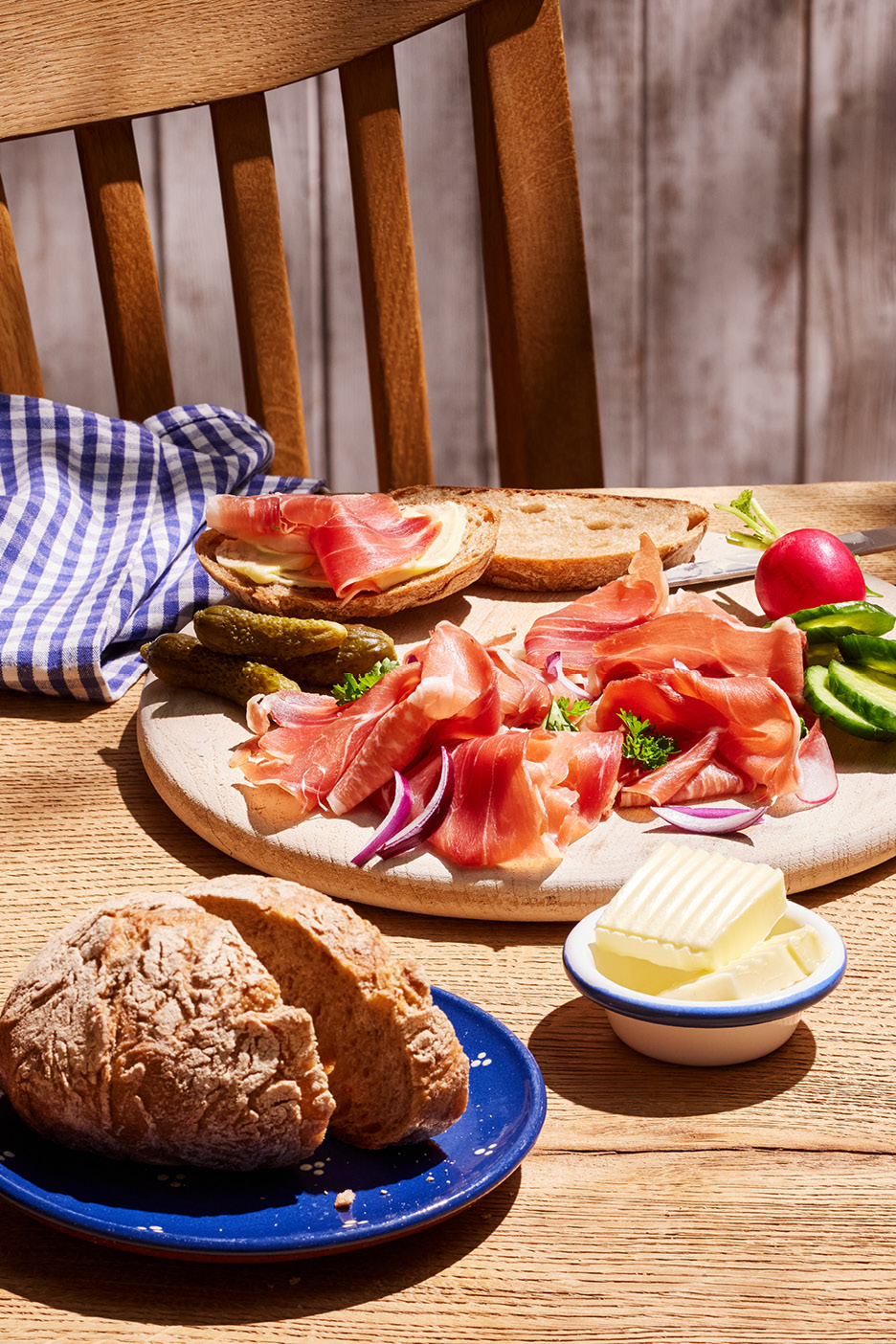 En rustik träskiva med skinka, bröd, smör, saltgurka och rädisor på ett träbord.