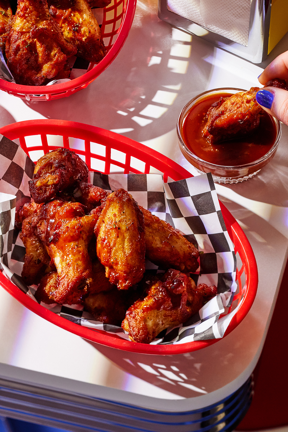 Friterade kycklingvingar i röda korgar med rutigt papper, en hand doppar en vinge i sås.