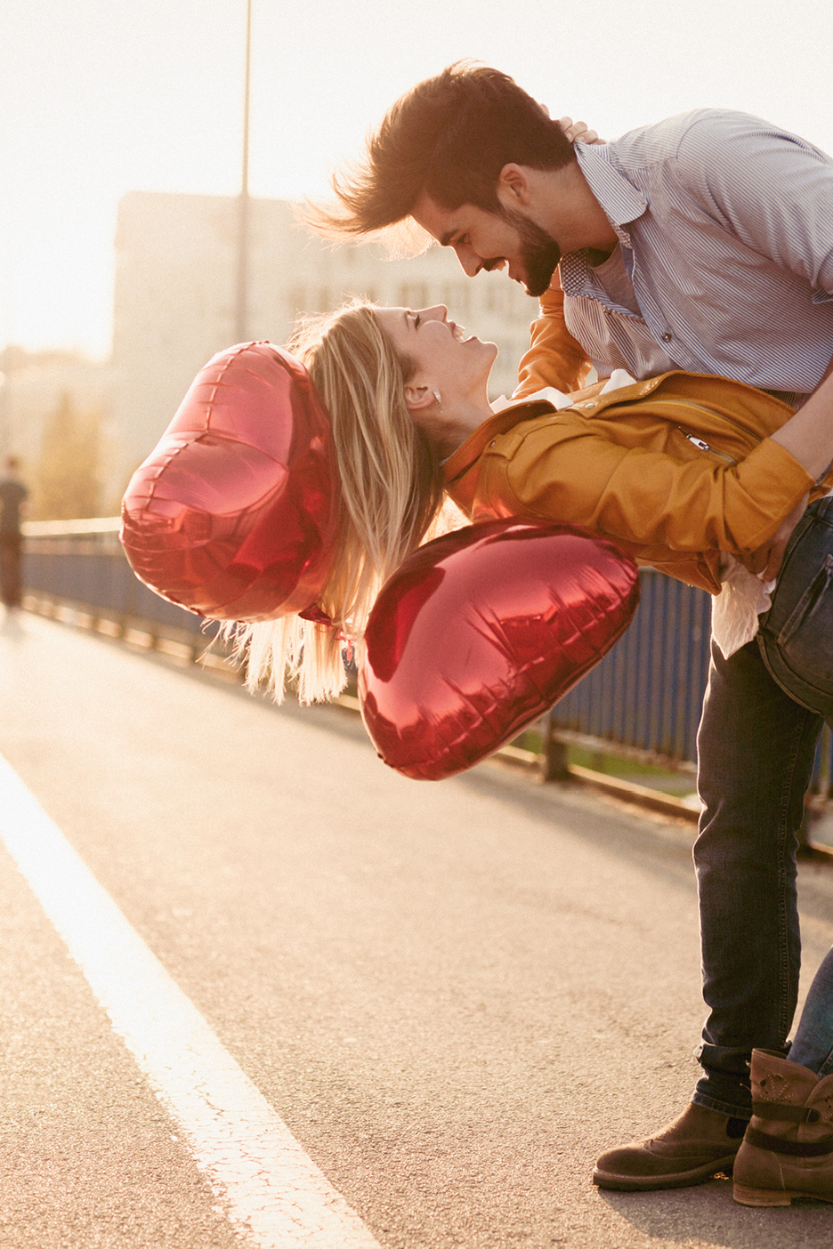 Ett par med hjärtformade ballonger på en bro, kvinnan lutar sig bakåt och mannen håller henne.