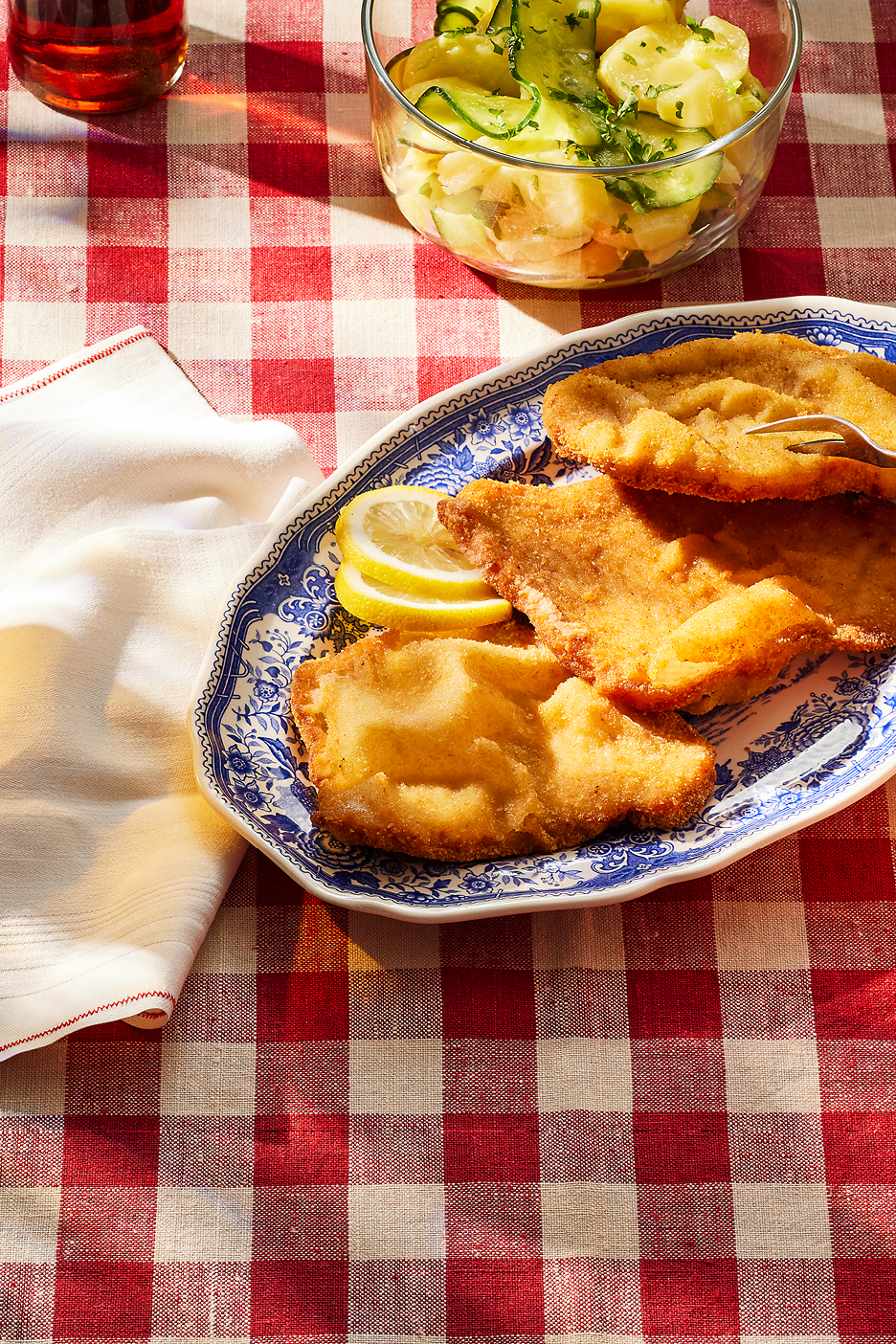 Friterad schnitzel med citronklyftor och potatis- och gurksallad på en rödvitrutig duk.