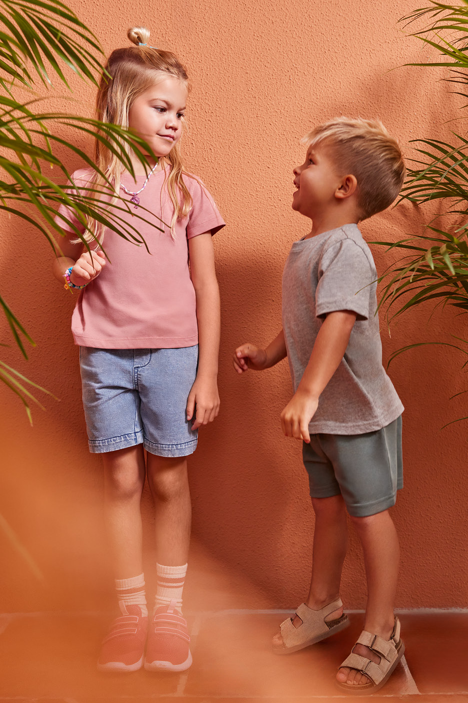 Två barn i sommarkläder, en flicka i rosa t-shirt och jeansshorts, en pojke i grå t-shirt och gröna shorts.