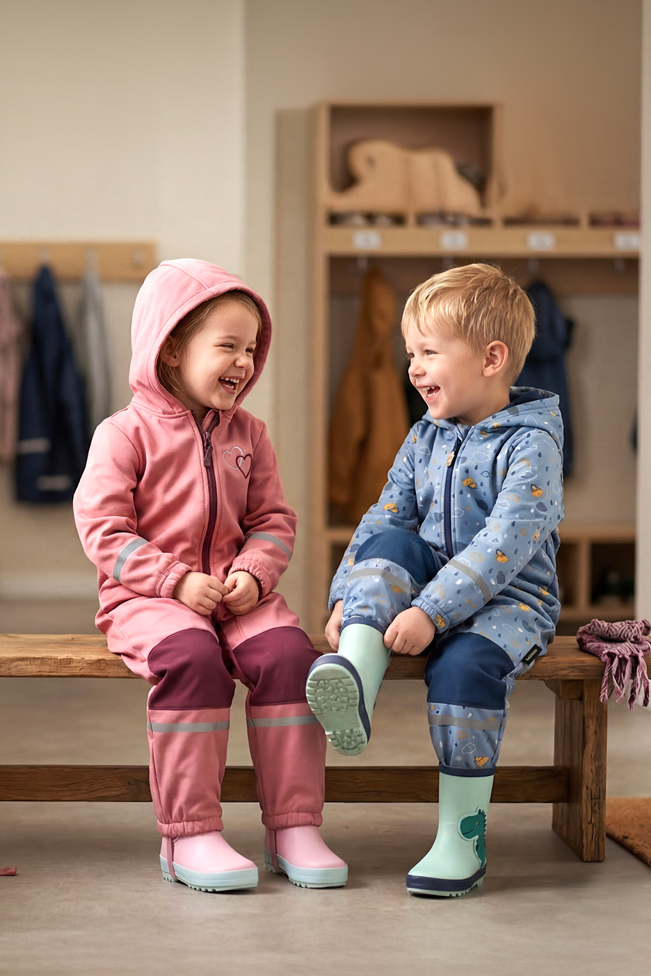 Två glada barn i rosa och blåa regnoveraller och gummistövlar sitter på en bänk.