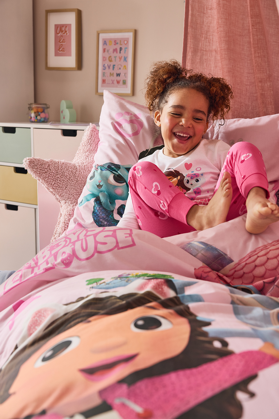 Glad flicka i rosa pyjamas med Gabbys dockhus-tema i en säng.