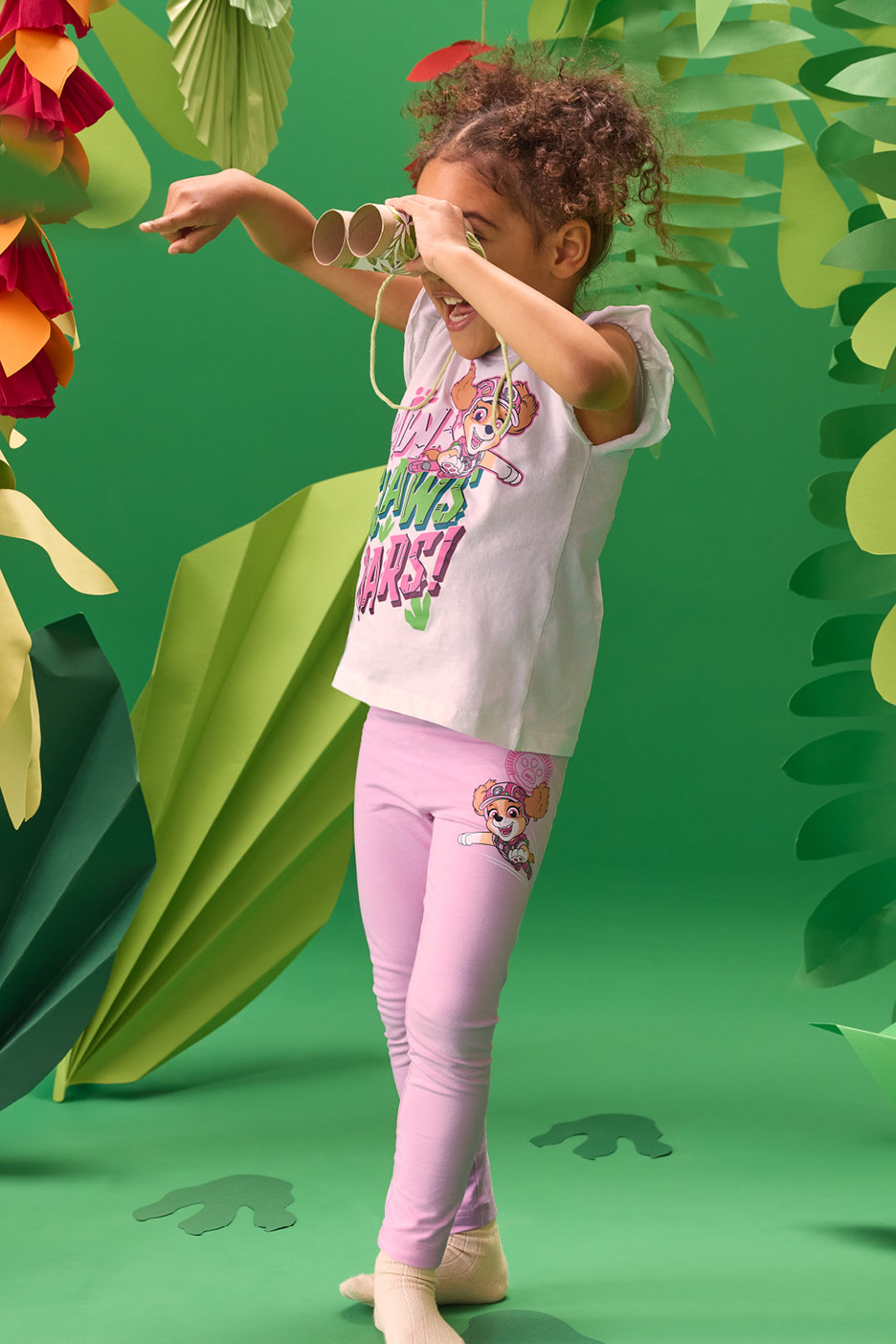 Flicka i vit t-shirt och rosa leggings leker med kikare av toarullar i en grön djungelmiljö.