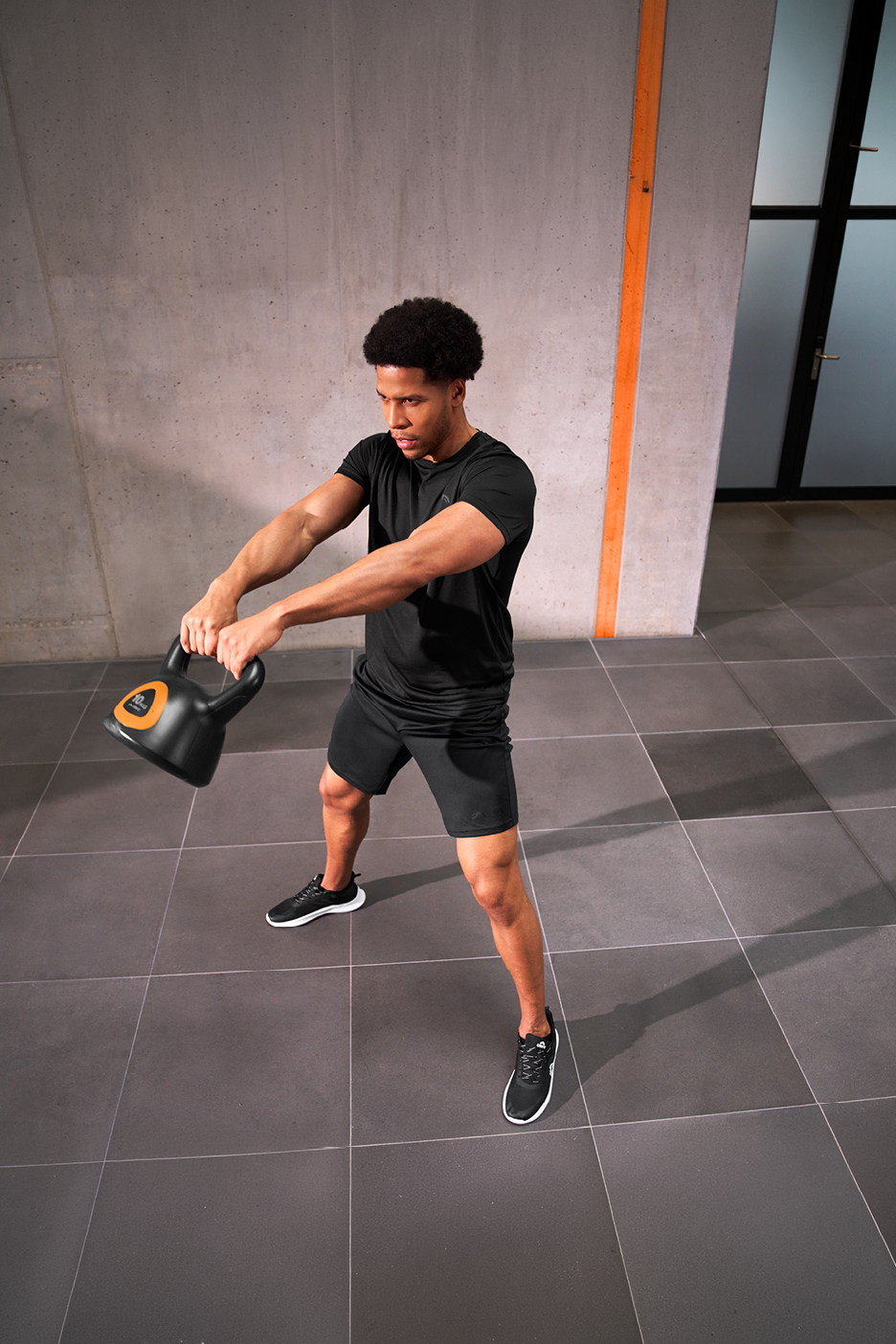 Man i träningskläder svingar en svart kettlebell med orange detaljer i ett gym.