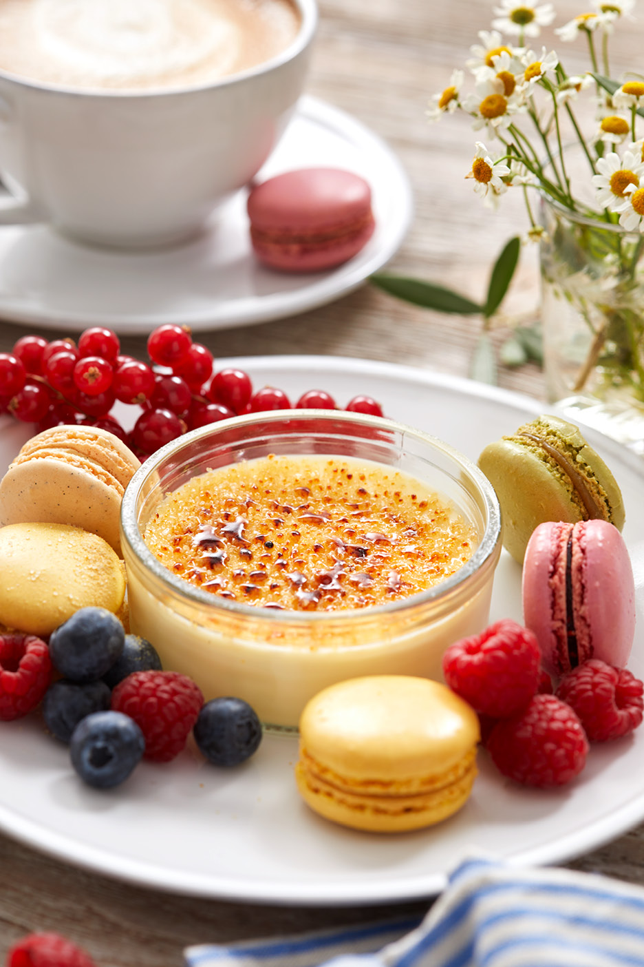 Crème brûlée med makaroner, bär och kaffe på ett träbord med blommor.