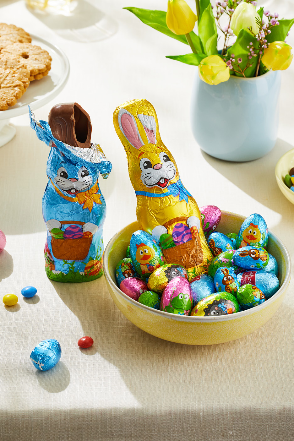 Chokladkaniner och påskägg i folie på ett bord med blommor och kakor.