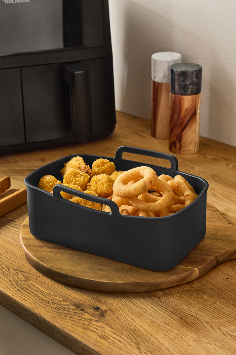 En svart airfryer med en korg fylld med lökringar och chicken nuggets på en träbänk.