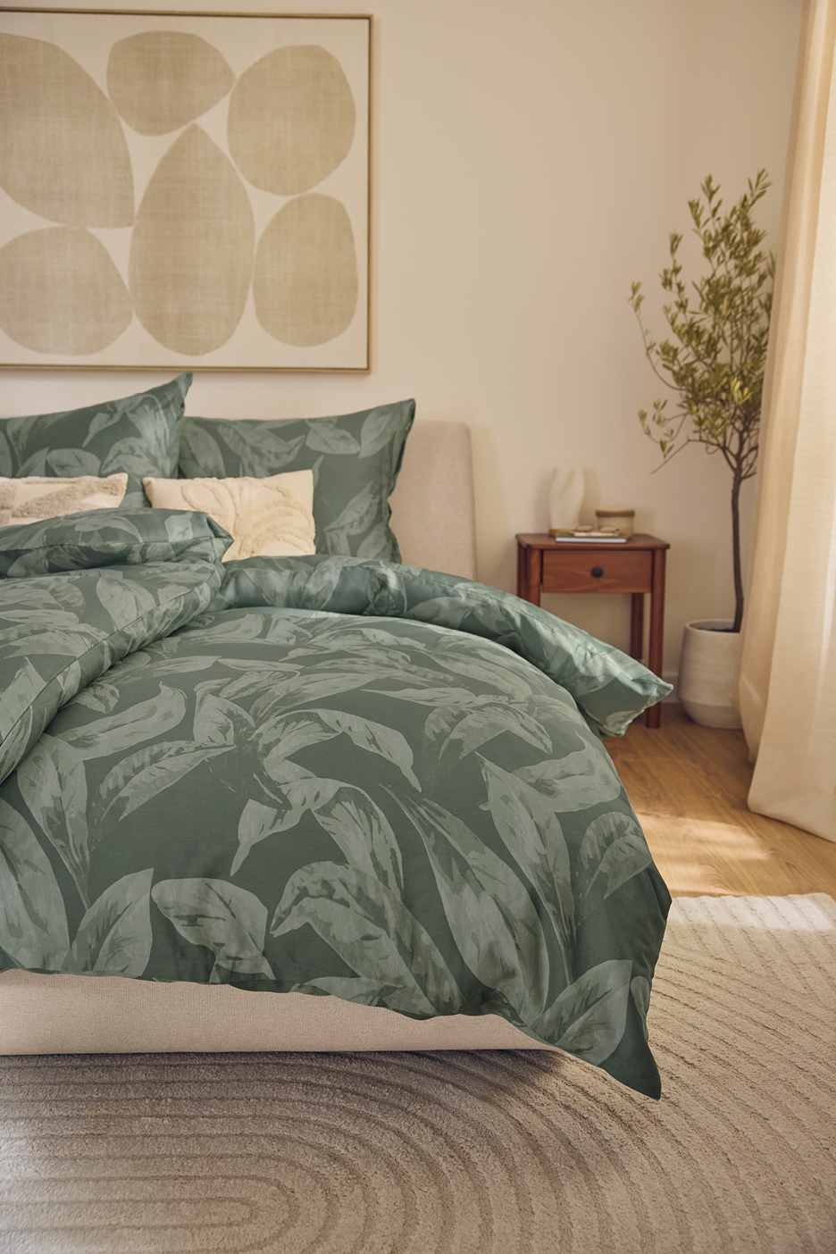 Sovrum med sängkläder i mörkgrönt med bladmönster, en sänggavel och en tavla.