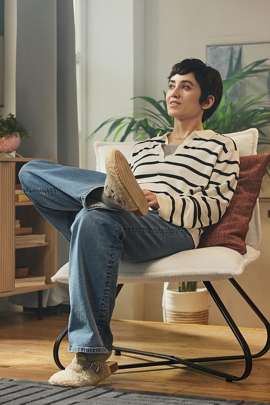 Kvinna i randig tröja och jeans sitter i en vit fåtölj med fårskinnstofflor.