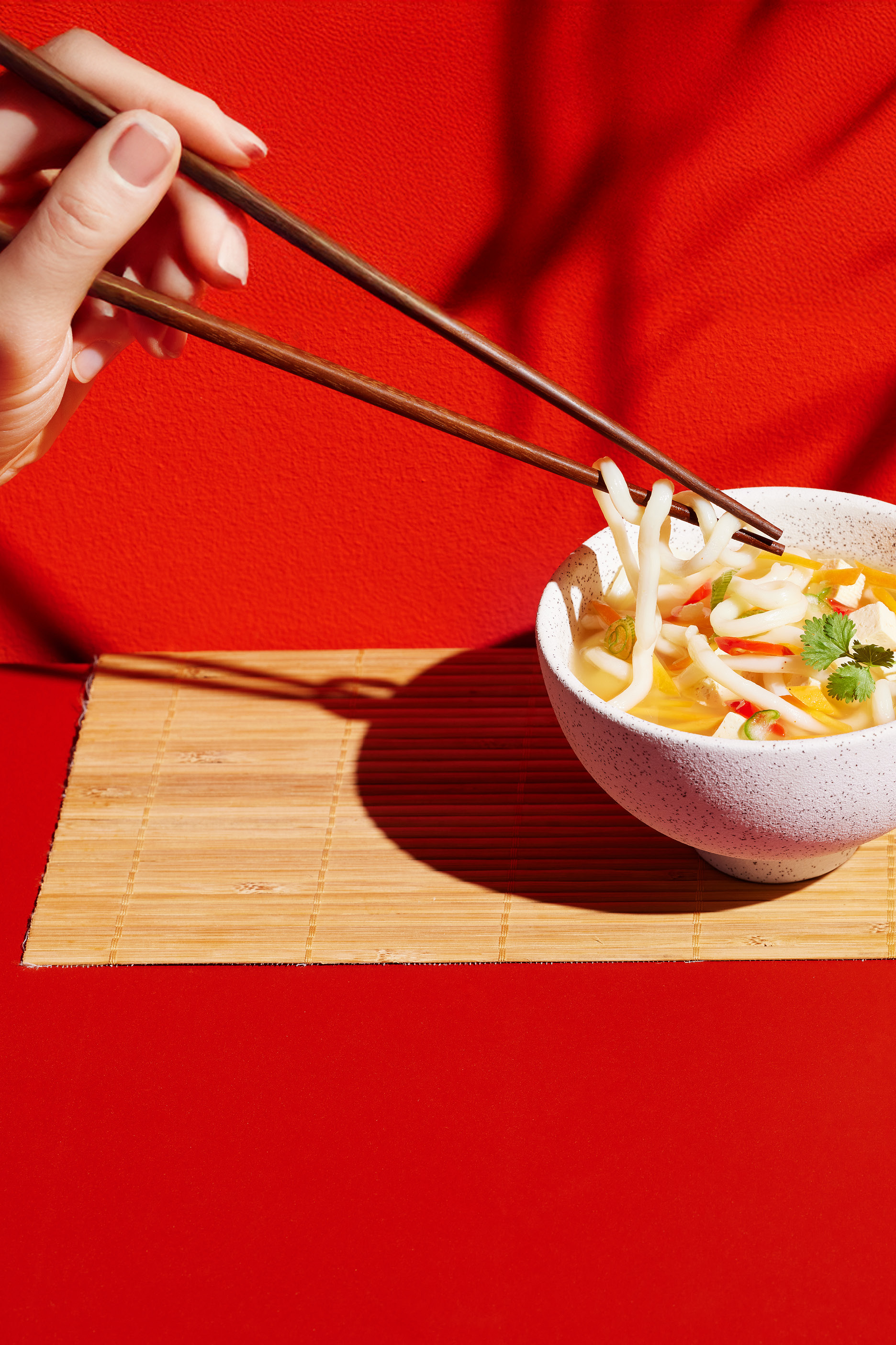 En hand med ätpinnar lyfter nudlar från en skål med nudelsoppa på en röd bakgrund.