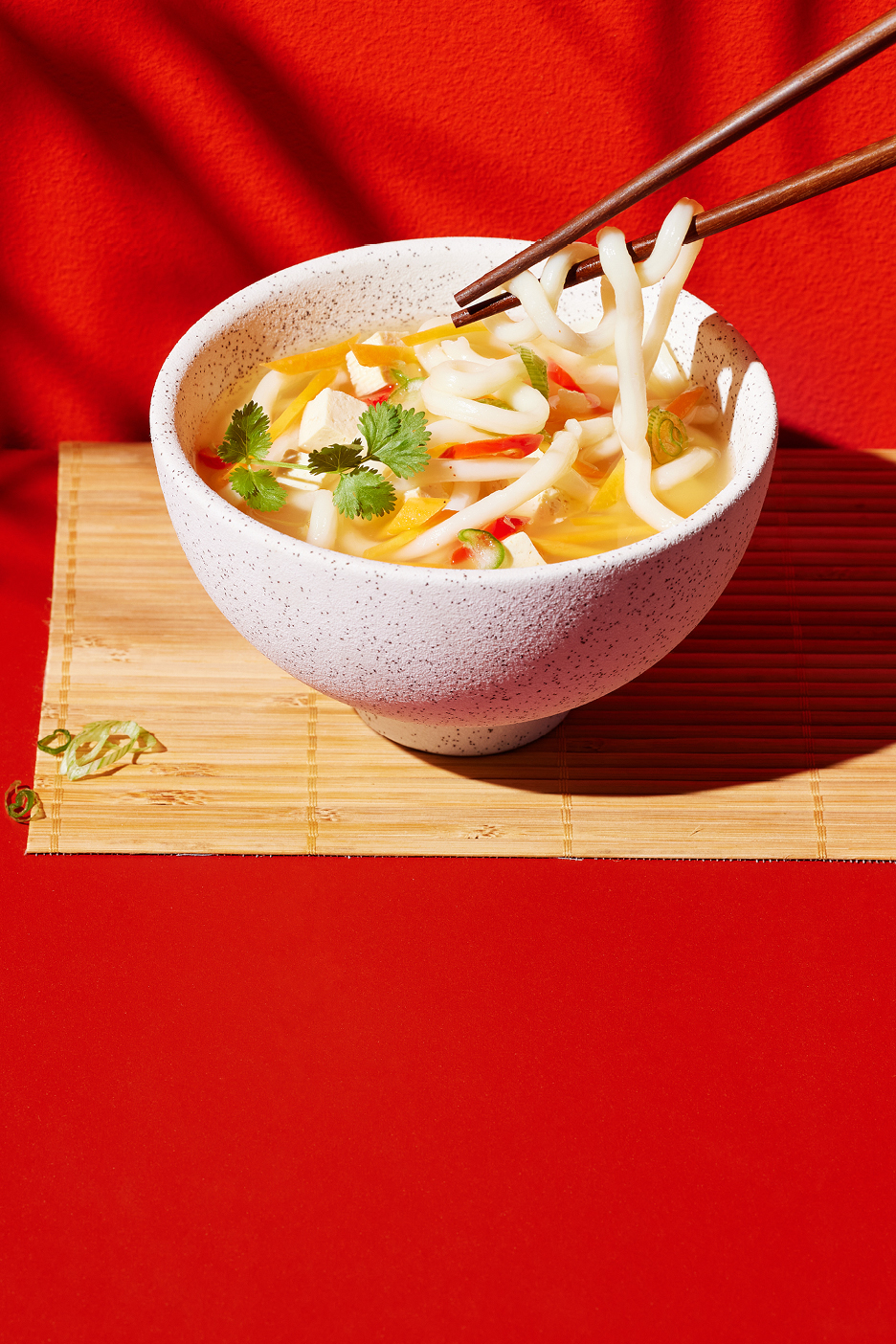 En skål med nudelsoppa med tofu och grönsaker, ätpinnar lyfter nudlar, på en bambumatta.