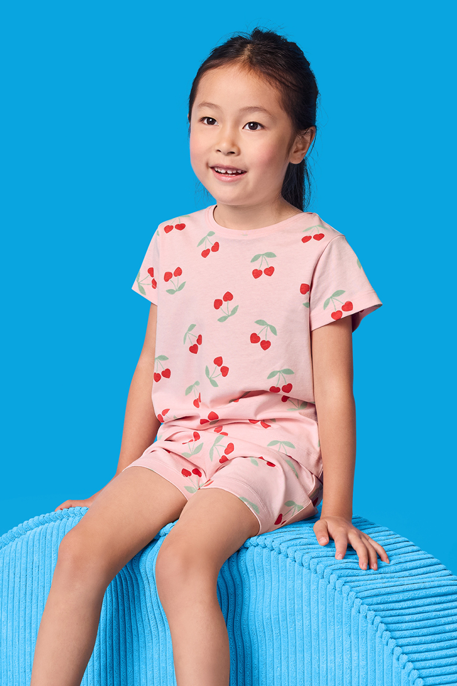 En glad flicka i rosa pyjamas med körsbärsmönster sitter på en blå kub.