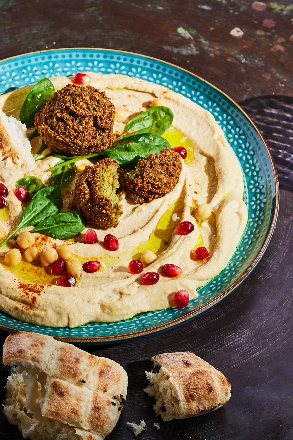 Hummus med falafel, granatäpple, kikärtor, spenat och bröd på en blå tallrik.