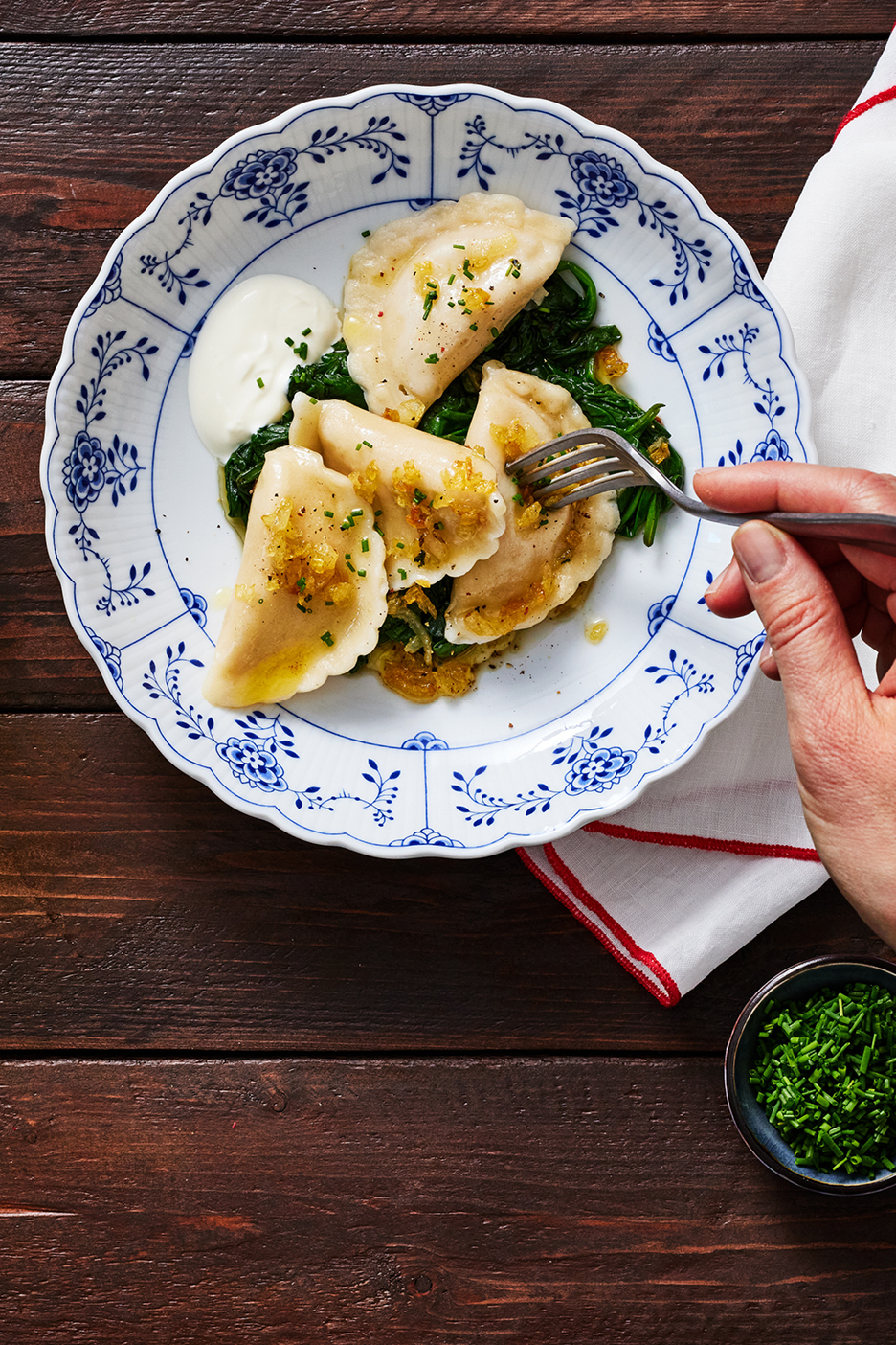 Pierogi med spenat, gräddfil och stekt lök på en tallrik, med en hand som håller en gaffel.