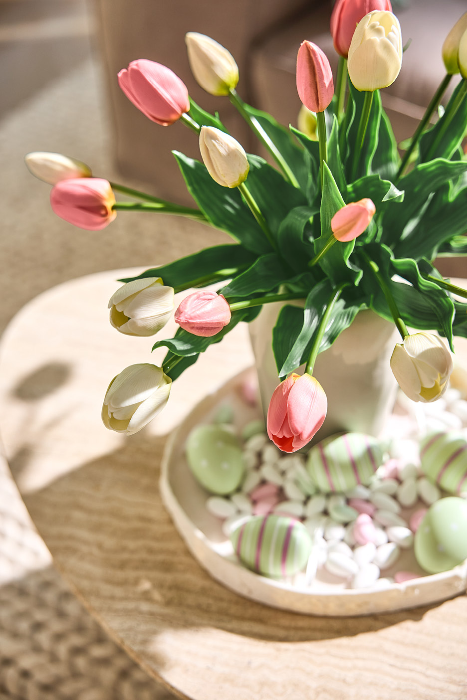 Rosa och vita tulpaner i vas med påskägg och godis på ett bord.