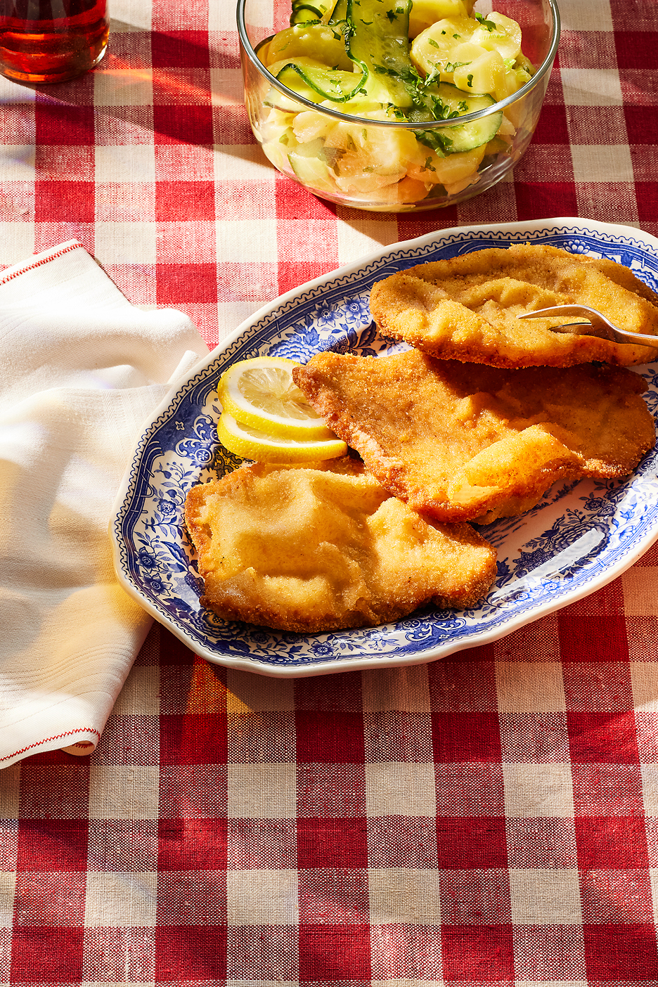 Panerad schnitzel med citron och potatis- och gurksallad på en rödvitrutig duk.