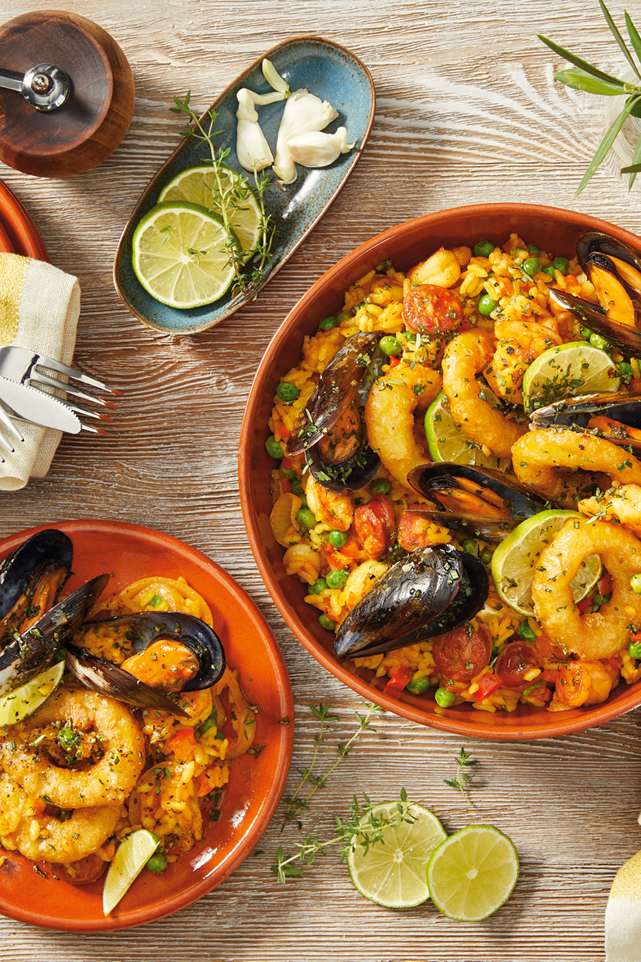 Paella med skaldjur, chorizo och limeklyftor i terrakottaskålar på träbord.