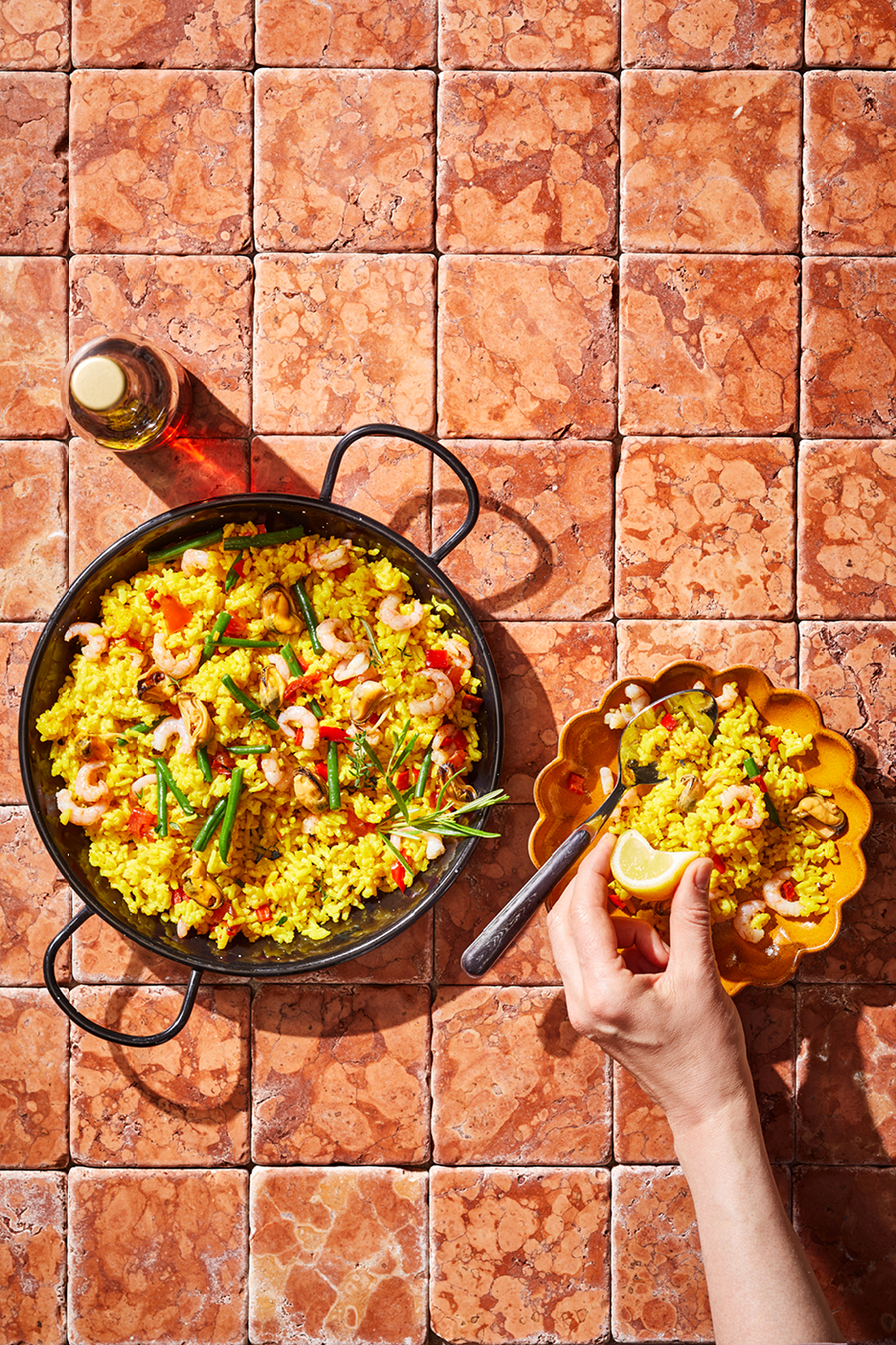 Paella med skaldjur och grönsaker i en stor panna och en portion på en tallrik med en hand som pressar citron.