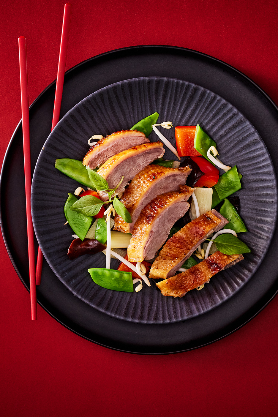 Anka med grönsaker och böngroddar på en svart tallrik med röda ätpinnar.