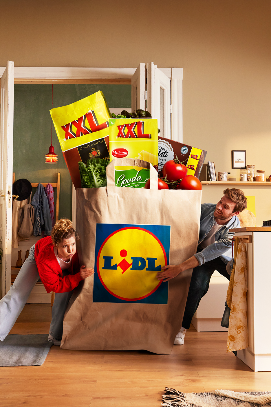 Man och kvinna bär en stor matkasse fylld med XXL-produkter, inklusive kaffe, ost och tomater.