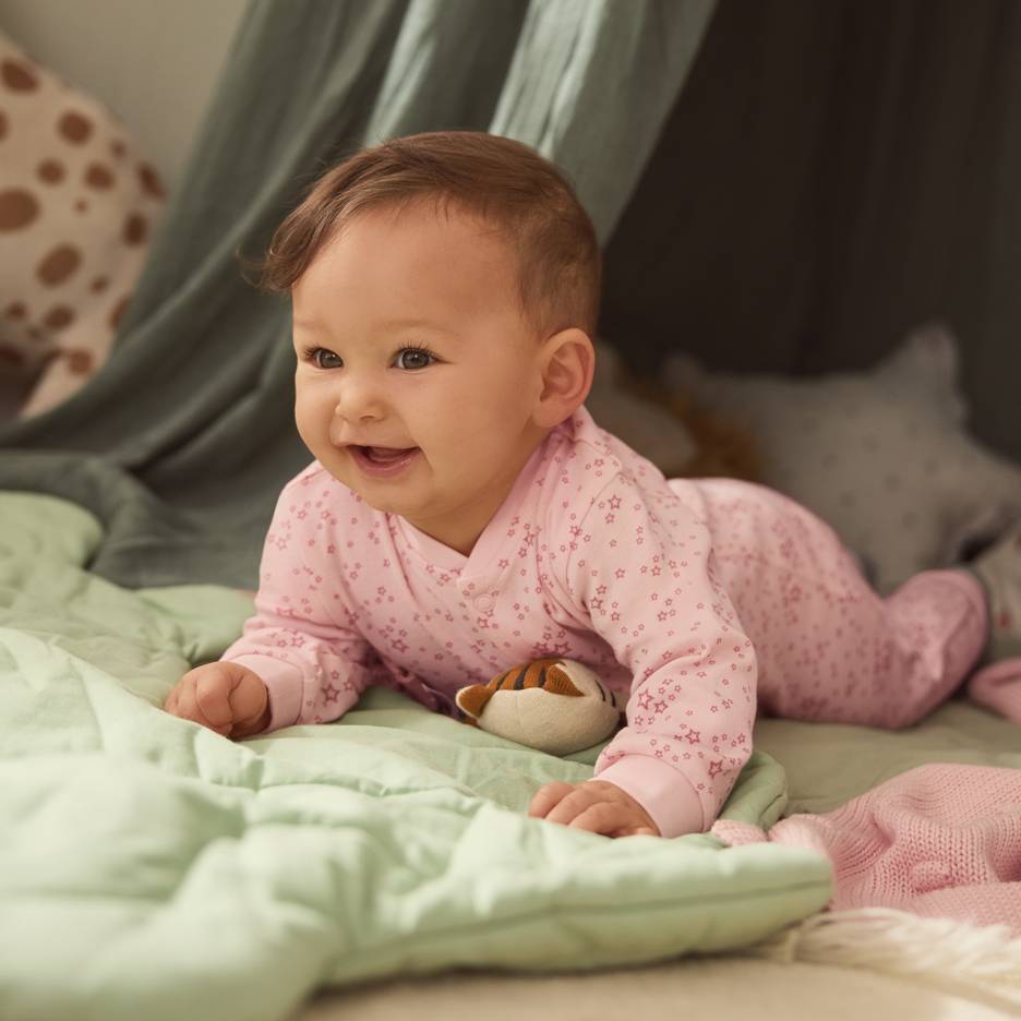 Baby i rosa stjärnpyjamas, liggande på en grön filt med en mjukdjur.