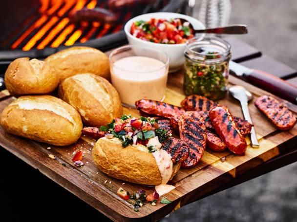 Grillade korvar och bröd med salsa och såser på en träskärbräda, med en grill i bakgrunden.
