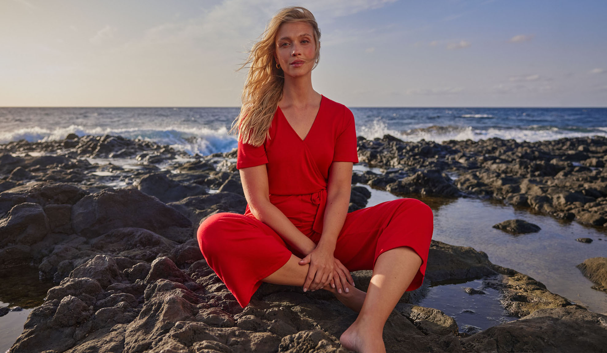 Kvinna i röd jumpsuit sitter på klippor vid havet.