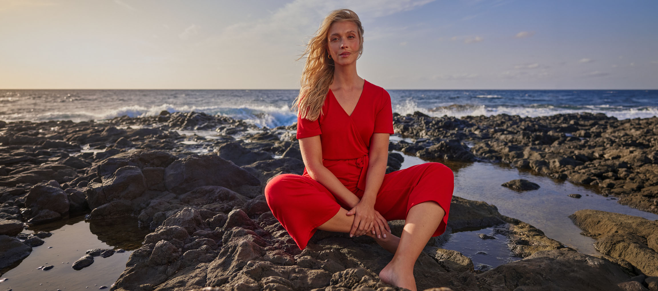 Kvinna i röd jumpsuit sitter på en stenig strand vid havet.