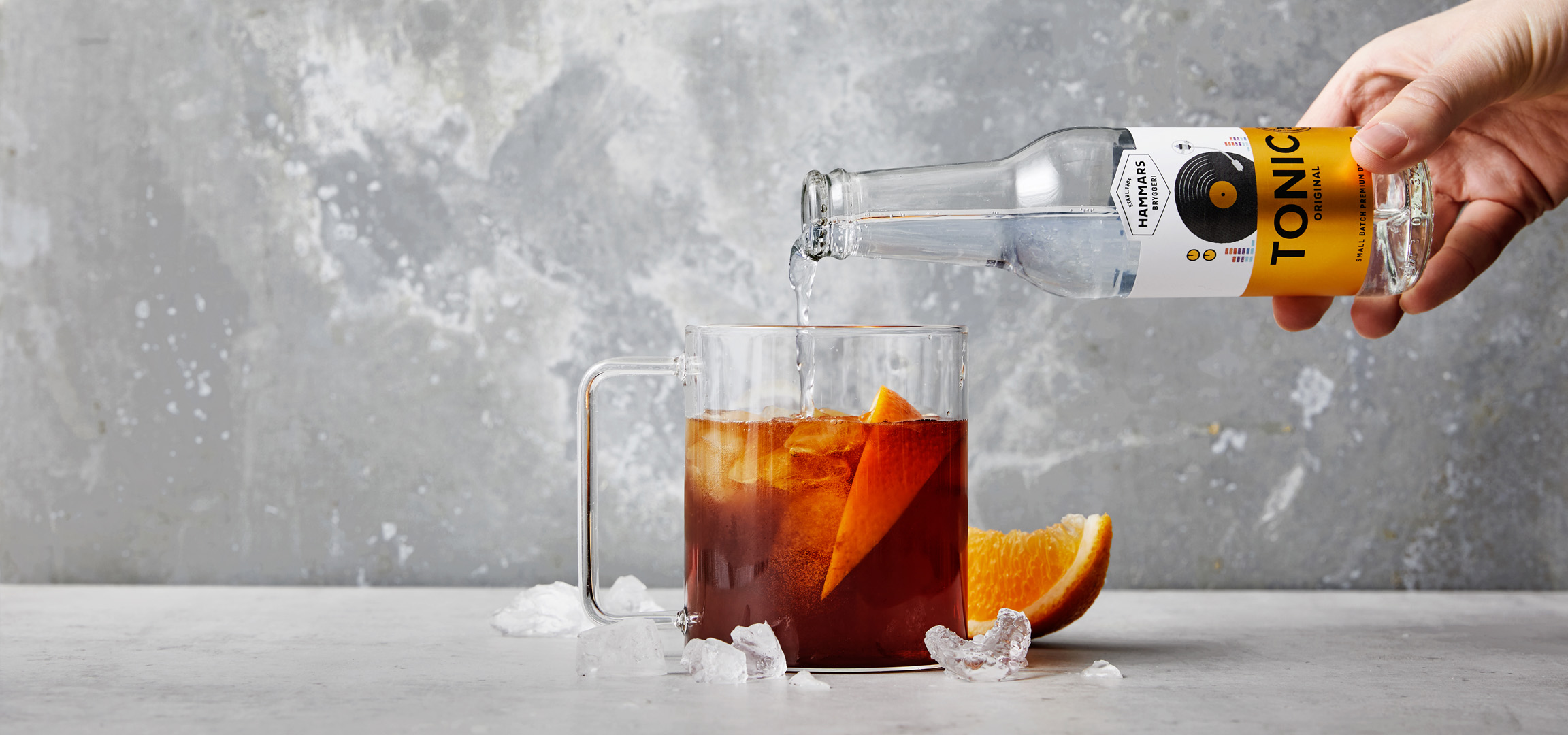 En hand häller Tonic Water i ett glas med is och apelsin.