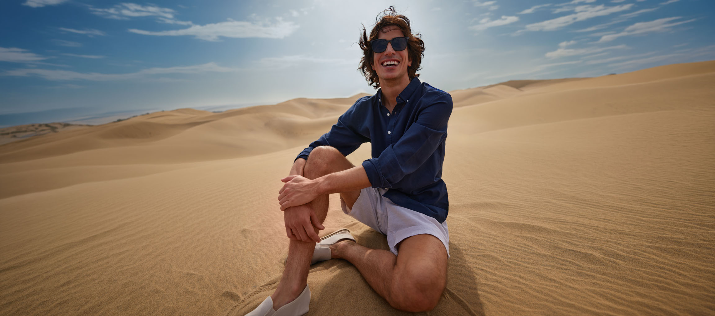 Man i mörkblå skjorta och shorts sitter leende i sanddynerna.