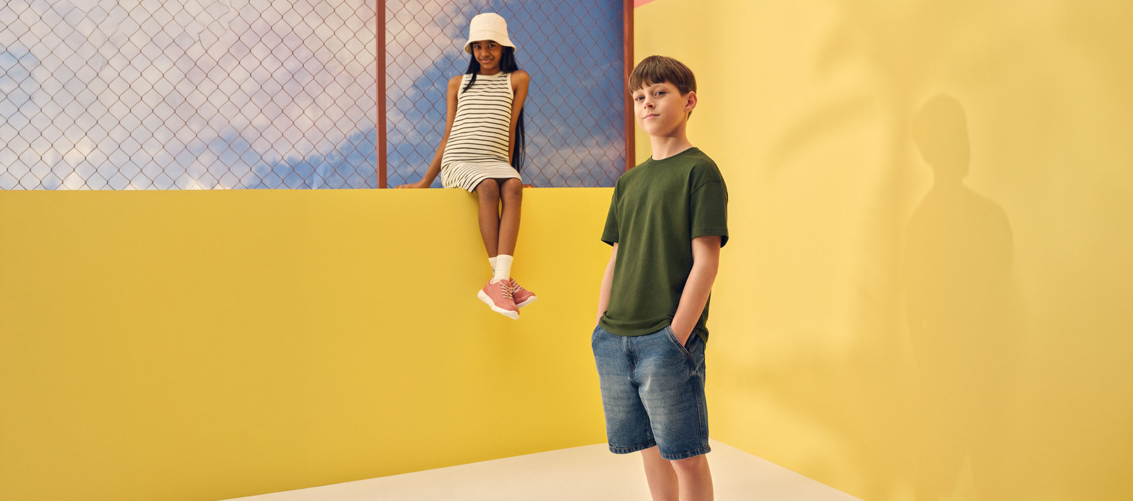 Två barn i avslappnade sommarkläder: en flicka i randig klänning och en pojke i grön t-shirt och jeansshorts.