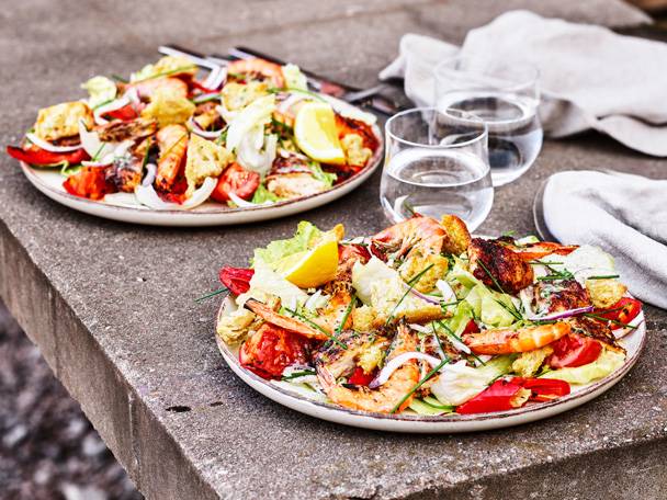 Två tallrikar med räk- och kycklingsallad med krutonger, grönsaker och citronklyftor på en stenbänk.