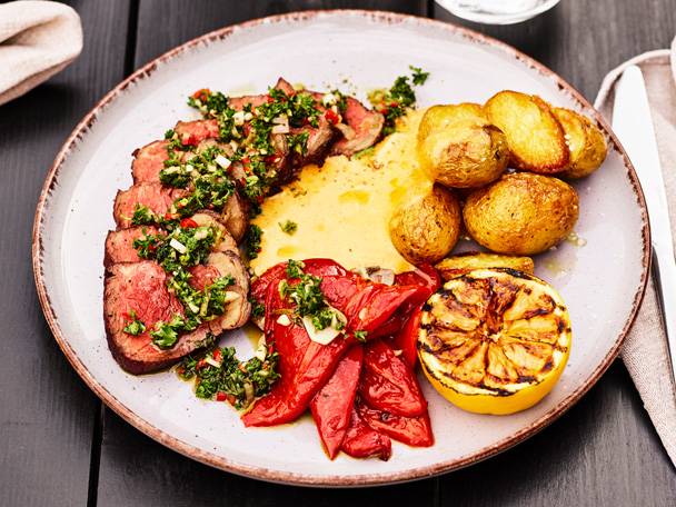 Skivad biff med chimichurri, grillad paprika, potatis och citron på en tallrik.