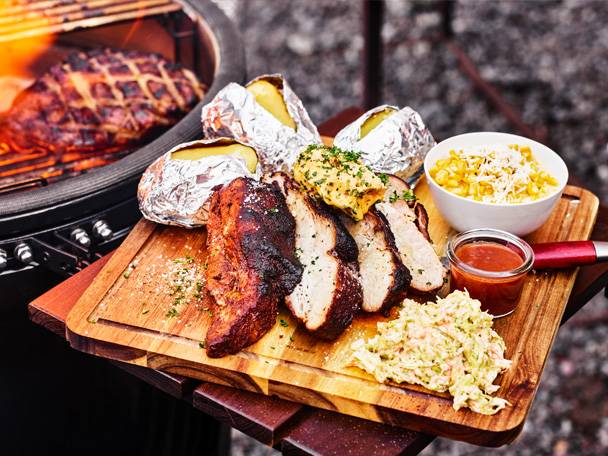 Grillad köttbit med bakpotatis, majs och coleslaw på en träskärbräda.