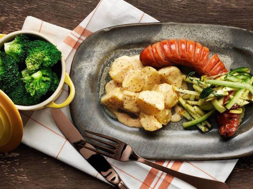 En tallrik med korv, potatis i sås, grönsaker och en skål med broccoli.